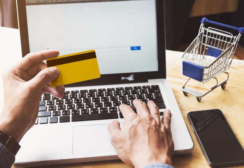 Zakupy online przy laptopie z kartą płatniczą i miniaturowym koszykiem, symbolizujące proces zakupowy w sklepie internetowym.